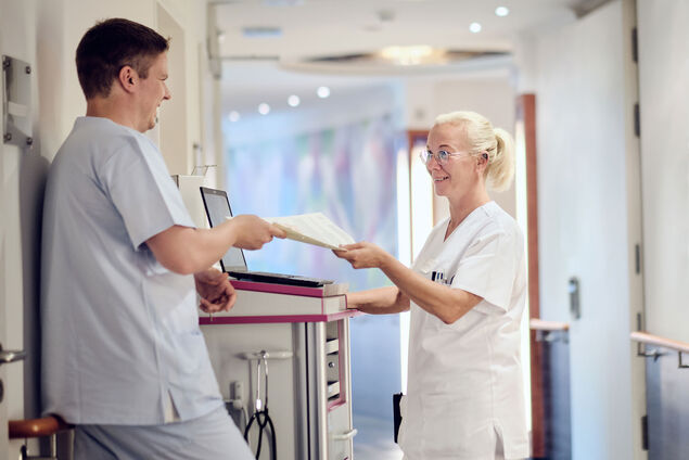 Zwei Pflegefachkräfte übergeben sich lächelnd auf dem Gang eine Patientenakte. 