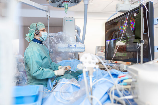 Arzt steht vor einem sehr großen Monitor mit einer 3D Animation des Herzens während er einen Katheter bei einem Patienten führt. 