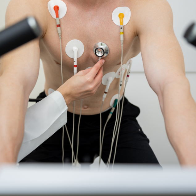 Ärztin bringt bei einem Mann EKG- Elektroden an der Brust an während er auf einem Fahrrad-ergometer sitzt. 