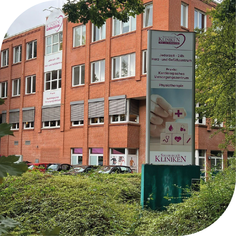 Rotes Backsteinhaus mit großer Klinik Beschiderung liegt komplett eingebettet ins Grüne an einer ruhigen Straße. 